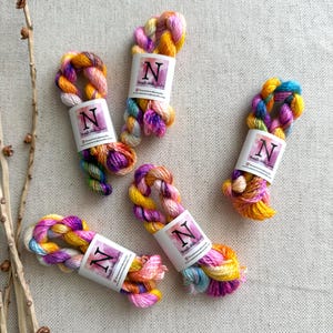 May include: Five skeins of hand-dyed embroidery floss featuring a variegated color palette of bright yellow, magenta, orange, and light blue. Each skein is wrapped in a white paper band with the letter N and the text Nanette Wake Studio.