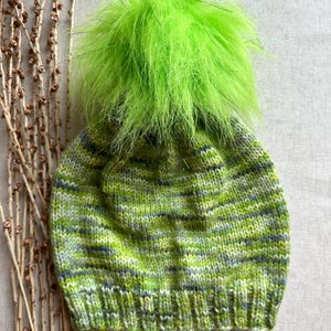 May include: A green knit hat with a green fuzzy pom pom. The hat has a striped pattern in shades of green, white, and gray.