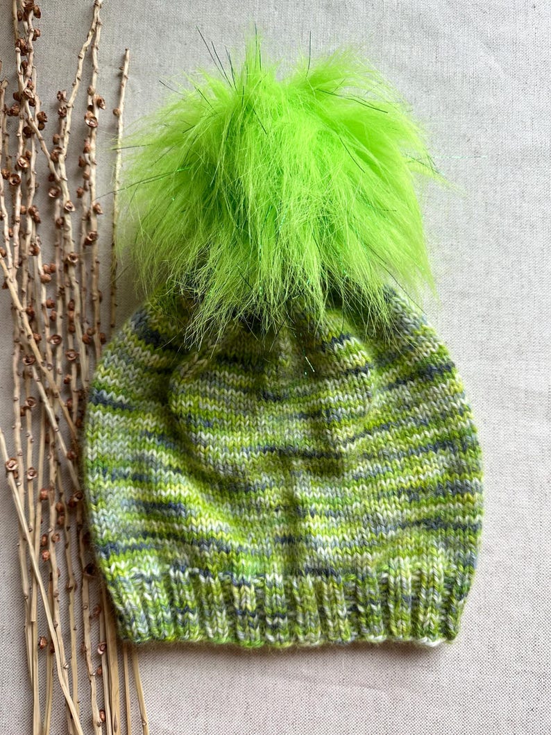 May include: A green knitted hat with a green fluffy pom pom. The hat has a striped pattern in shades of green, white and grey.