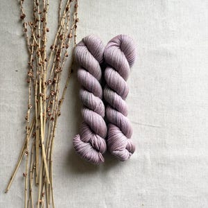 May include: Two skeins of hand-dyed yarn in shades of lavender and gray sit on a neutral linen surface. Dried branches with small brown buds are arranged to the left of the yarn. The yarn is tightly wound, showing the texture and color variations.