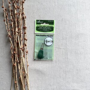 May include: A package of Clover Yarn Threader, a green and white tool for threading yarn, with the text "Makes threading yarns easy!". The package is on a neutral background with dried branches.