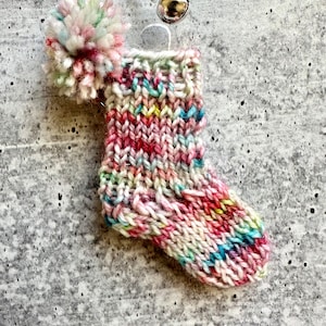 May include: A small, hand-knitted Christmas stocking pendant with a colourful pom-pom. The stocking is mainly white, pink, and red, with blue, yellow, and green accents. It hangs from a silver chain with a small, round charm.