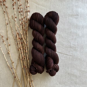 May include: Two skeins of dark brown yarn, wound into balls, on a beige fabric background.