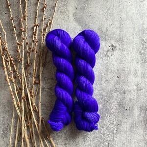 May include: Two skeins of vibrant purple yarn are displayed on a textured gray surface, with dried branches on the left. The yarn is tightly wound, showcasing a rich, saturated color. This image is ideal for showcasing knitting or crochet supplies.