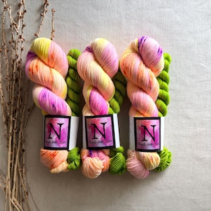 May include: Three skeins of hand-dyed yarn featuring a variegated color palette of bright yellow, soft peach, and vibrant magenta, paired with smaller skeins of solid lime green yarn. Each main skein has a white label with a black letter N.