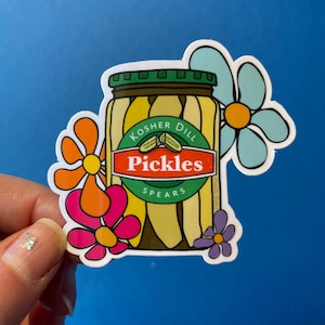 以下が含まれることがあります： コッシャーディルピクルス「コッシャーディルピクルススピアーズ」と書かれたラベルのついた瓶のステッカー。瓶はカラフルな花に囲まれています。