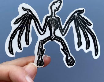 Op de afbeelding: Zwart-witte sticker van een vleermuis skelet met uitgespreide vleugels. De sticker is op een witte achtergrond.