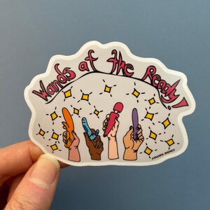 May include: A sticker with the text "Wands of the Ready!" in pink lettering. The sticker features a white background with four hands holding different colored wands, surrounded by yellow stars.