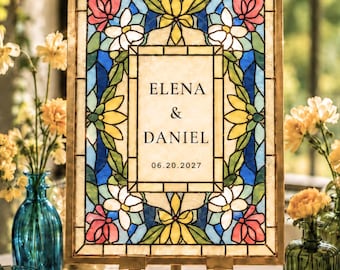 Puede incluir: Un letrero rectangular con un borde de vidrieras con patrones florales amarillos, azules, blancos y rosas. El panel central es de color crema con el texto negro ELENA & DANIEL y 06.20.2027. El letrero se exhibe en un pequeño caballete de madera.