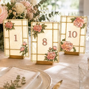 May include: Three rectangular table number signs featuring a cream-colored background with a grid pattern, pink peony illustrations, and the numbers 1, 8, and 10 in a serif font. Each sign is held upright by a small, rectangular gold metal stand.