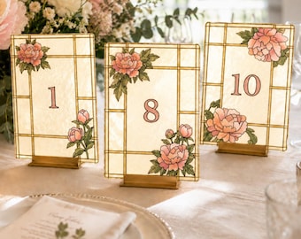 Puede incluir: Tres letreros rectangulares de números de mesa con fondo color crema, patrón de cuadrícula, ilustraciones de peonías rosas y los números 1, 8 y 10 en fuente serif. Cada letrero se sostiene mediante un pequeño soporte rectangular de metal dorado.