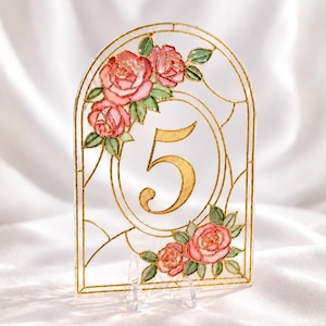 May include: An arched table number sign featuring the number 5 in gold, framed by a gold-lined stained glass style border with hand-painted pink roses and green leaves. The sign is supported by a small, clear acrylic stand for upright display.