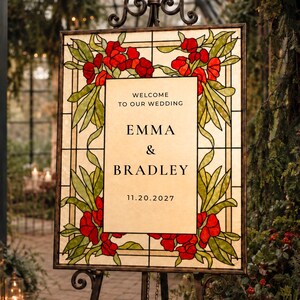 Puede incluir: Un letrero de bienvenida de boda rectangular con un borde de vidrieras de flores rojas y hojas verdes. El panel central color crema tiene texto negro que dice: WELCOME TO OUR WEDDING, EMMA & BRADLEY, 11.20.2027, sobre un soporte de metal.