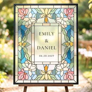 May include: A rectangular sign with a black frame featuring a stained glass pattern of pink, yellow, blue, and green floral motifs. The centre panel displays the text EMILY & DANIEL and the date 06.20.2027 in a black serif font on a light background.