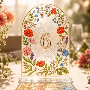 May include: An arched glass table number sign featuring the number 6 in gold, surrounded by a colorful painted floral wreath with red, pink, blue, and white flowers and green leaves, set on a clear rectangular base with a textured glass surface.