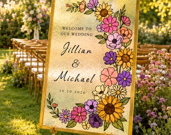Puede incluir: Un letrero de bienvenida de boda rectangular con marco dorado sobre un trípode. El letrero presenta un fondo dorado con un borde floral de girasoles y flores moradas, rosas y naranjas. El texto dice: Welcome to our wedding, Jillian & Michael, 10.20.2026.