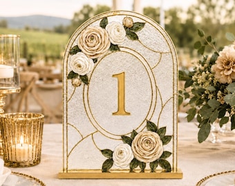 Puede incluir: Un letrero de mesa arqueado con el número 1 en dorado, sobre un fondo blanco texturizado con un marco dorado. El diseño incluye detalles florales pintados con rosas de color crema y hojas verdes en un estilo de vidriera.