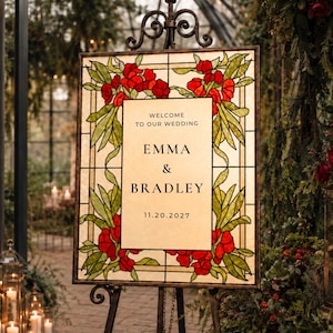 May include: A rectangular wedding welcome sign featuring a stained glass border with red flowers and green leaves. The center panel is cream-colored with black text reading: WELCOME TO OUR WEDDING, EMMA & BRADLEY, 11.20.2027, displayed on a metal stand.