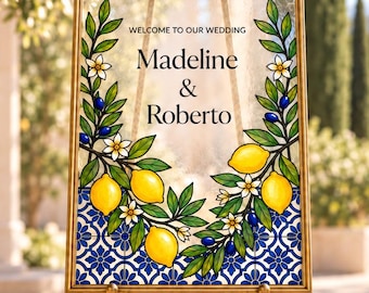 Puede incluir: Un letrero de bienvenida de boda rectangular en un marco dorado sobre un caballete dorado. El letrero presenta el texto "WELCOME TO OUR WEDDING Madeline & Roberto" con un borde decorativo de limones amarillos, hojas verdes, flores blancas y patrones de azulejos azules.