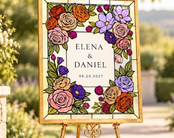 Puede incluir: Un letrero rectangular con marco dorado sobre un caballete de metal dorado. El letrero presenta un borde de vidrieras con rosas y flores coloridas que rodea un panel central blanco con el texto ELENA & DANIEL y la fecha 06.20.2027 en negro.