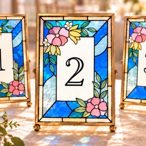 May include: Three rectangular table number frames with a stained glass border featuring blue geometric patterns, pink flowers, and green leaves. Each frame has a white center panel displaying the black numbers 1, 2, and 3 respectively.