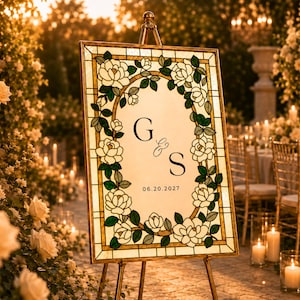 May include: A rectangular wedding sign on a metal easel featuring a stained glass-style border of white roses and green leaves. The center is white with the black serif letters G and S, an ampersand, and the date 06.20.2027 printed in black.