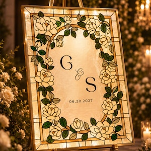 May include: A rectangular sign featuring a stained glass border with white flowers and green leaves surrounding a central panel. The panel displays the letters G & S in a serif font with the date 06.20.2027 printed in black below the letters.