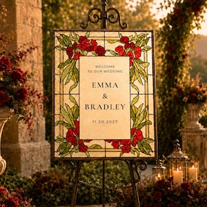 May include: A rectangular wedding sign featuring a stained glass border with red flowers and green leaves. The center panel is cream-colored with black text reading: WELCOME TO OUR WEDDING, EMMA & BRADLEY, 11.20.2027. The sign is displayed on a black metal easel.