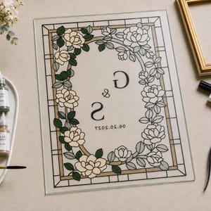 May include: A rectangular glass panel featuring a stained-glass style border of cream and green roses. The center displays the letters G and S with the date 06.20.2027. Surrounding items include glass paint bottles, a brush, and a black liner.