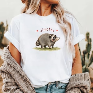 Może przedstawiać: Biały t-shirt z kr&oacute;tkim rękawem z grafiką przedstawiającą szarego szopa pracza stojącego na kępce zielonej trawy. Nad szopem znajduje się napis "jimothy" wydrukowany ciemnoczerwoną czcionką, otoczony dwiema małymi czerwonymi ikonami serc.