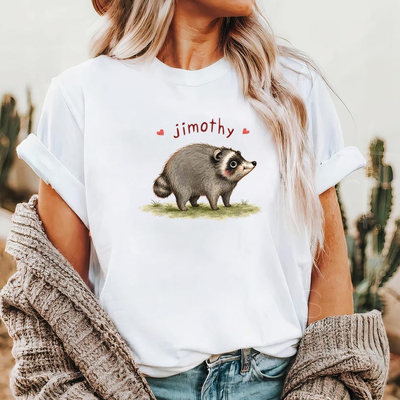Może przedstawiać: Biały t-shirt z kr&oacute;tkim rękawem z grafiką przedstawiającą szarego szopa pracza stojącego na kępce zielonej trawy. Nad szopem znajduje się napis "jimothy" wydrukowany ciemnoczerwoną czcionką, otoczony dwiema małymi czerwonymi ikonami serc.