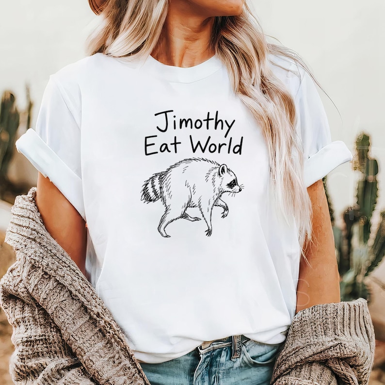Może przedstawiać: Biały t-shirt z kr&oacute;tkim rękawem z czarnym rysunkiem liniowym szopa pracza i napisem "Jimothy Eat World" wydrukowanym prostą, ciemną czcionką na klatce piersiowej. Koszulka ma okrągły dekolt i jest noszona z brązowym, teksturowanym kardiganem.