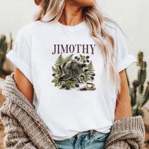 Może przedstawiać: Biały t-shirt z nadrukiem graficznym przedstawiającym szopa pracza w otoczeniu zielonych liści paproci i ciemnych jeżyn. Pod szopem znajduje się mała filiżanka kawy na spodku. Napis JIMOTHY jest wydrukowany ciemnofioletową czcionką nad szopem.