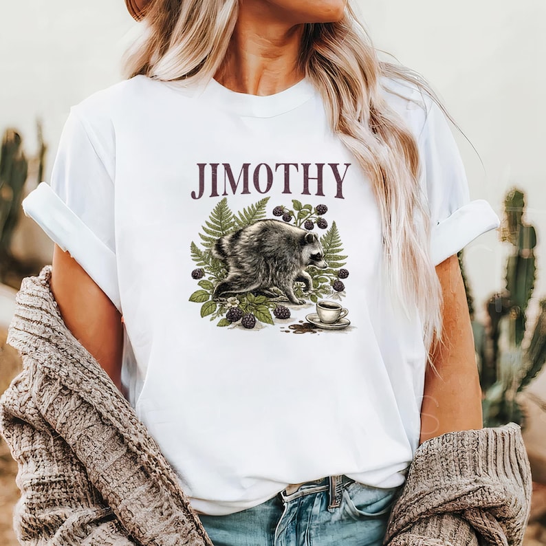 Może przedstawiać: Biały t-shirt z nadrukiem graficznym przedstawiającym szopa pracza w otoczeniu zielonych liści paproci i ciemnych jeżyn. Pod szopem znajduje się mała filiżanka kawy na spodku. Napis JIMOTHY jest wydrukowany ciemnofioletową czcionką nad szopem.
