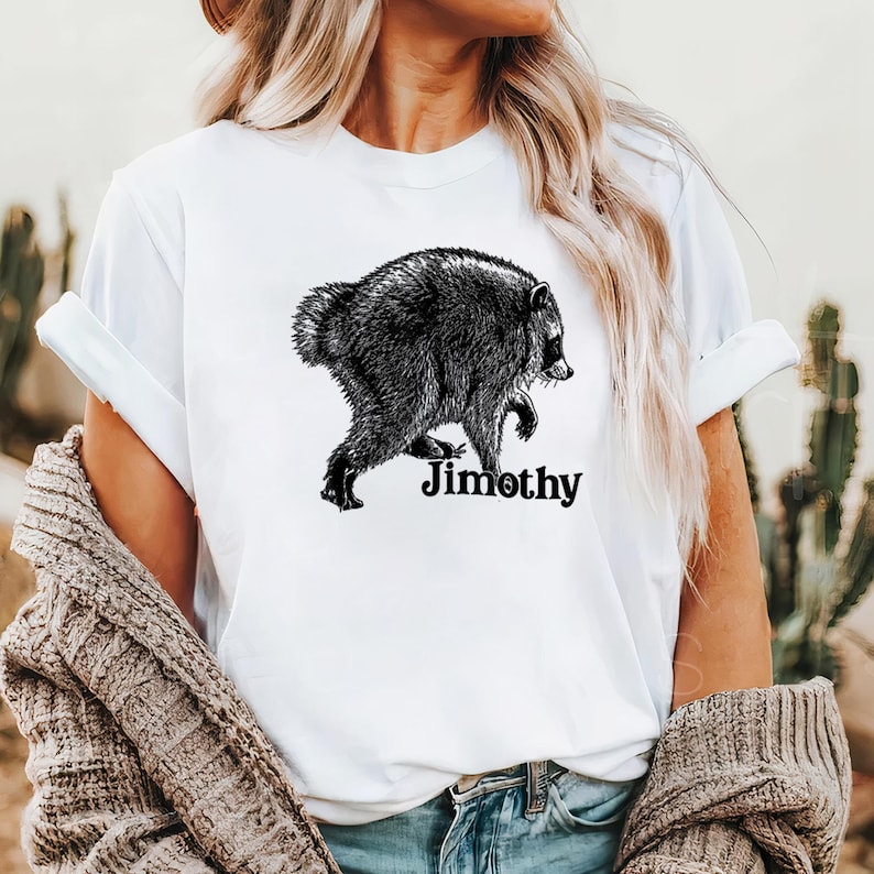 Może przedstawiać: Biały t-shirt z kr&oacute;tkim rękawem z czarno-białą ilustracją szopa pracza z profilu. Pod szopem znajduje się napis Jimothy wykonany pogrubioną, czarną czcionką szeryfową. Koszulka posiada okrągły dekolt i podwinięte rękawy.