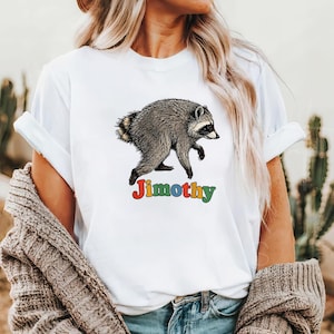 Może przedstawiać: Biały t-shirt z kr&oacute;tkim rękawem z ilustracją szaro-czarnego szopa pracza w pozycji marszu. Pod szopem znajduje się napis "Jimothy" wykonany wielokolorową, zaokrągloną czcionką z czerwonymi, ż&oacute;łtymi, niebieskimi i zielonymi literami.