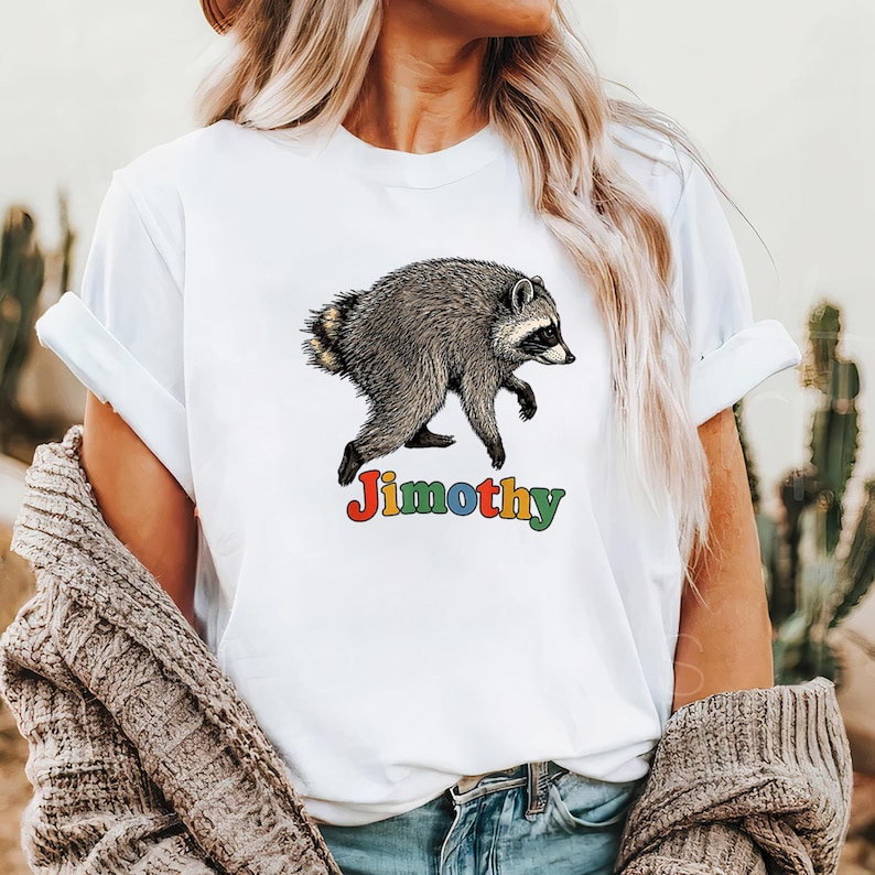 Może przedstawiać: Biały t-shirt z kr&oacute;tkim rękawem z ilustracją szaro-czarnego szopa pracza w pozycji marszu. Pod szopem znajduje się napis "Jimothy" wykonany wielokolorową, zaokrągloną czcionką z czerwonymi, ż&oacute;łtymi, niebieskimi i zielonymi literami.