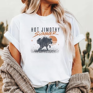 Może przedstawiać: Biały t-shirt z kr&oacute;tkim rękawem z grafiką przedstawiającą szopa pracza idącego przez trawę na tle zachodzącego słońca. Tekst nad grafiką brzmi HOT JIMOTHY SUMMER czarnymi i pomarańczowymi literami, z dodatkiem małych ikon gwiazdek.