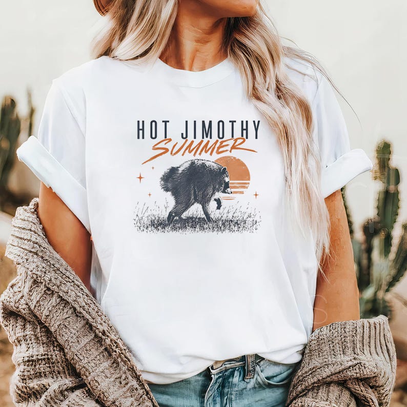 Może przedstawiać: Biały t-shirt z kr&oacute;tkim rękawem z grafiką przedstawiającą szopa pracza idącego przez trawę na tle zachodzącego słońca. Tekst nad grafiką brzmi HOT JIMOTHY SUMMER czarnymi i pomarańczowymi literami, z dodatkiem małych ikon gwiazdek.