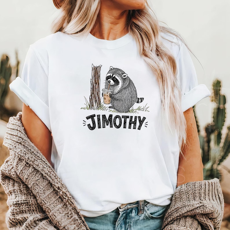Może przedstawiać: Biały t-shirt z grafiką przedstawiającą szopa pracza stojącego obok pnia drzewa i trzymającego plastikowy kubek ze słomką. Pod szopem znajduje się napis JIMOTHY wykonany pogrubionymi, czarnymi, odręcznie narysowanymi wielkimi literami.