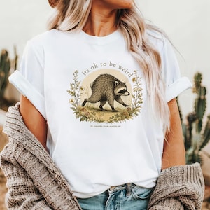 Może przedstawiać: Biały t-shirt z grafiką szopa pracza idącego wewnątrz kolistego wieńca kwiatowego. Tekst "its ok to be weird" jest wygięty w łuk nad szopem, a "jimothy from seattle" jest wydrukowany małymi literami na dole całego wzoru.