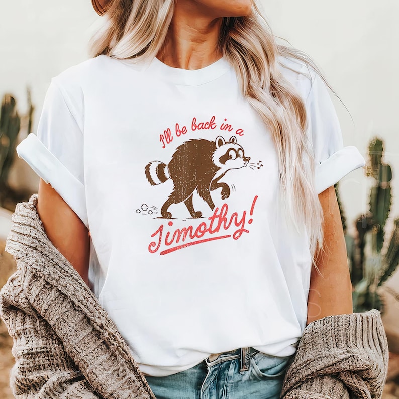 Może przedstawiać: Biały t-shirt z kr&oacute;tkim rękawem, grafiką brązowego szopa pracza i czerwonym napisem "I'll be back in a Jimothy!". Szop jest przedstawiony w pozycji marszowej, z nutką przy pysku i śladem kurzu za sobą.
