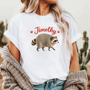 Może przedstawiać: Biały t-shirt z kr&oacute;tkim rękawem z grafiką przedstawiającą idącego brązowo-szarego szopa pracza. Nad szopem znajduje się napis "Jimothy" wykonany czerwoną kursywą, otoczony dwoma małymi czerwonymi sercami po obu stronach tekstu.