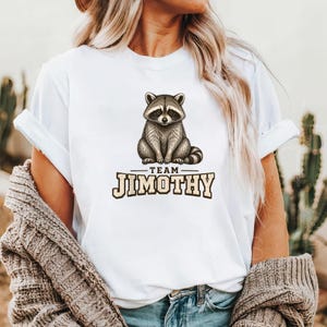 Może przedstawiać: Biały t-shirt z kr&oacute;tkim rękawem z grafiką przedstawiającą siedzącego szopa pracza. Poniżej szopa znajduje się napis "TEAM JIMOTHY" pogrubioną czcionką blokową. Koszulka ma okrągły dekolt i jest noszona z brązowym, teksturowanym kardiganem.