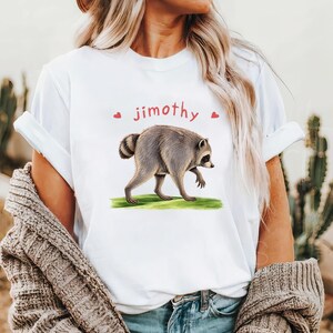 Może przedstawiać: Biały t-shirt z grafiką przedstawiającą szopa pracza idącego po kawałku zielonej trawy. Nad szopem znajduje się napis "jimothy" w kolorze czerwonym, otoczony dwoma małymi czerwonymi sercami. Koszulka ma kr&oacute;tkie rękawy i okrągły dekolt.