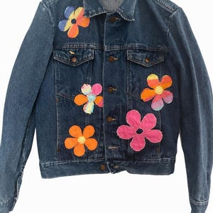 May include: A blue denim jacket adorned with colorful, embroidered flower appliqués. The flowers are in shades of orange, pink, yellow, and blue. The jacket has a classic design with button closures and pockets, offering a vintage, handcrafted aesthetic.