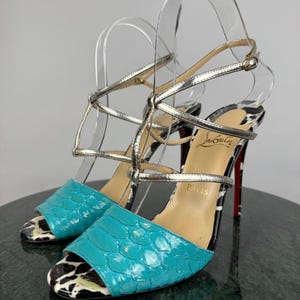 May include: A pair of high-heeled sandals with a turquoise snakeskin pattern on the toe strap. The sandals have silver straps and a black and white zebra print on the heel. The soles are red.