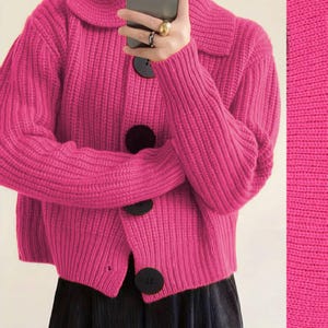 Puede incluir: Un cárdigan rosa brillante de punto grueso con un cuello grande y tres botones negros de gran tamaño. Las mangas son largas y la tela tiene una textura acanalada. El cárdigan es de color liso.