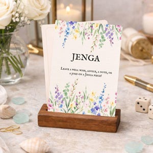 Puede incluir: Un juego de Jenga con diseño floral de acuarela. Las cartas se muestran en un soporte de madera. El texto en las cartas dice "JENGA" y "¡Deja un buen deseo, un consejo, una nota o una broma en una pieza de Jenga!" También se ven dados, un bolígrafo y velas.
