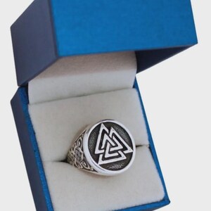 Puede incluir: Un anillo de plata con una cara circular que presenta un diseño geométrico audaz, presentado en una caja de joyería azul. La cara del anillo muestra un símbolo estilizado prominente. La caja está abierta, revelando el anillo.